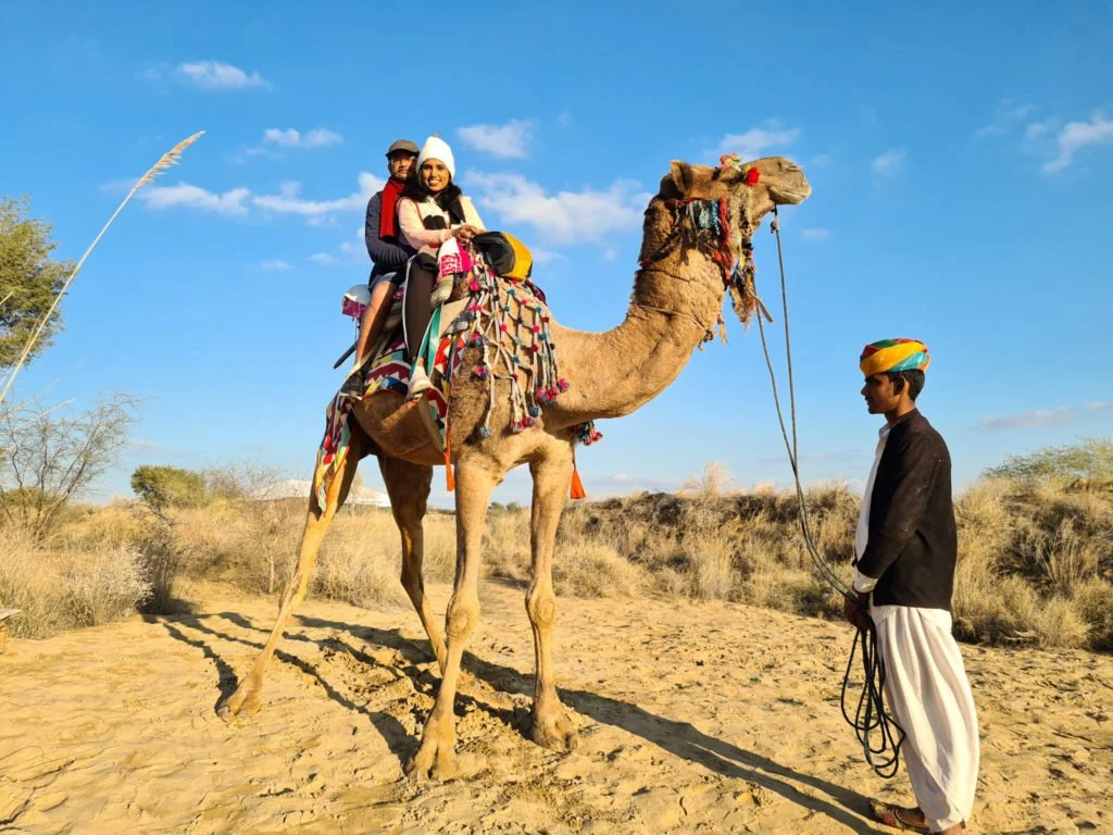 Roopesh and Kiki from RooKiExplorers sitting on a camel as the camel's owner holds the reigns.