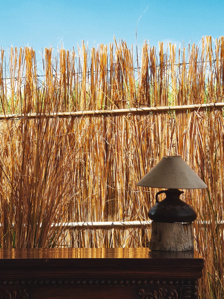 A lamp on a table with a fence made of straw behind the table.