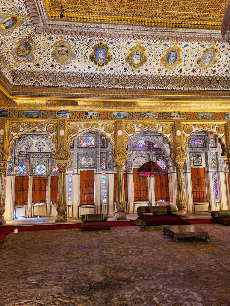 A room filled with paintings in Mehrangarh fort. The ceiling and pillars have gold on them.