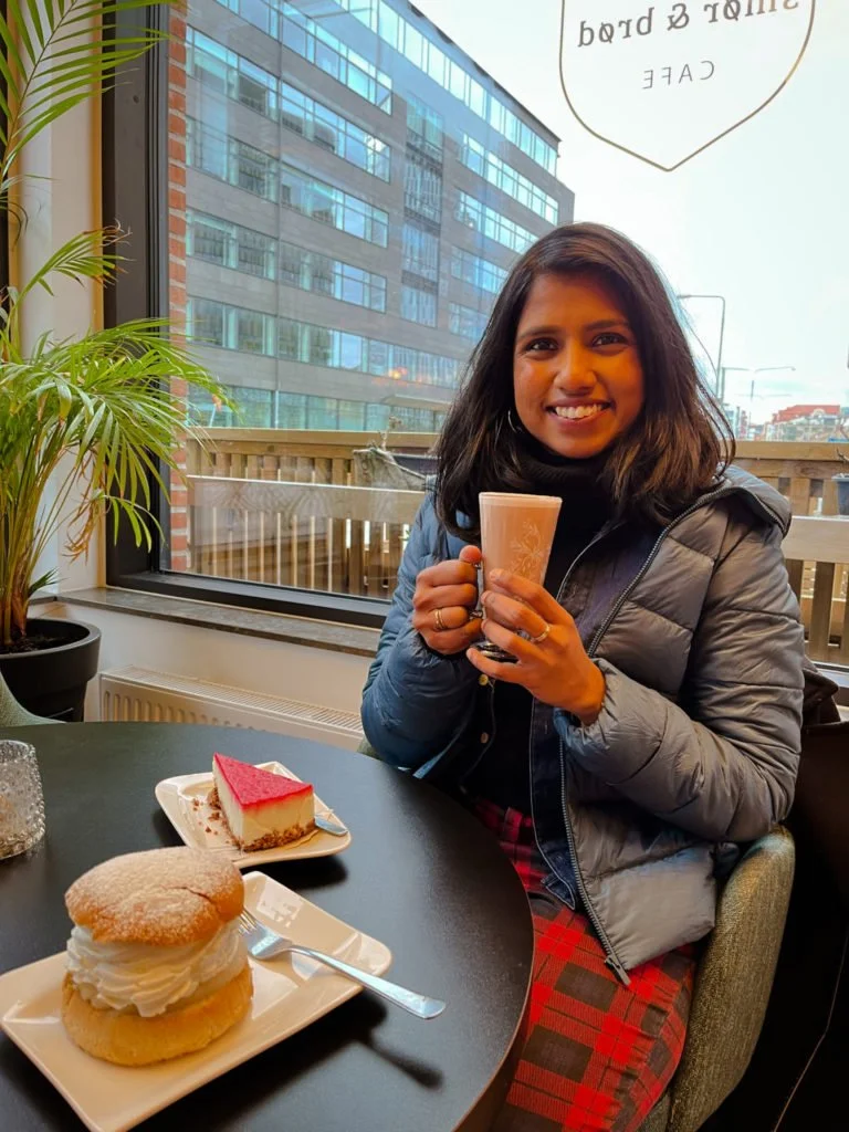 Kiki from RooKiExplorers holding a hot chocolate in a cafe in Gustav Adolfs Torg. There is strawberry cheesecake and semla on the small dining table.