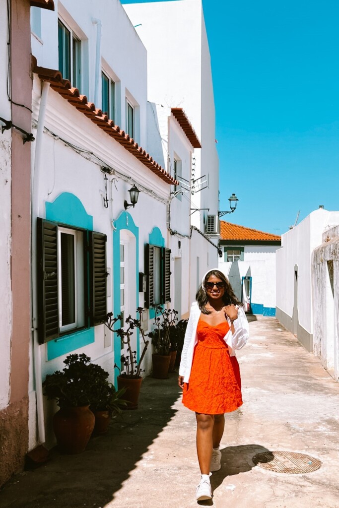 Kiki from RooKiExplorers posing in a street with blue and white colored houses in Ferragudo, Algarve.