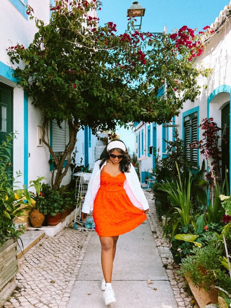 Kiki from RooKiExplorers strolling in a street with a bougainvillea tree and blue and white colored houses in Ferragudo, Algarve.