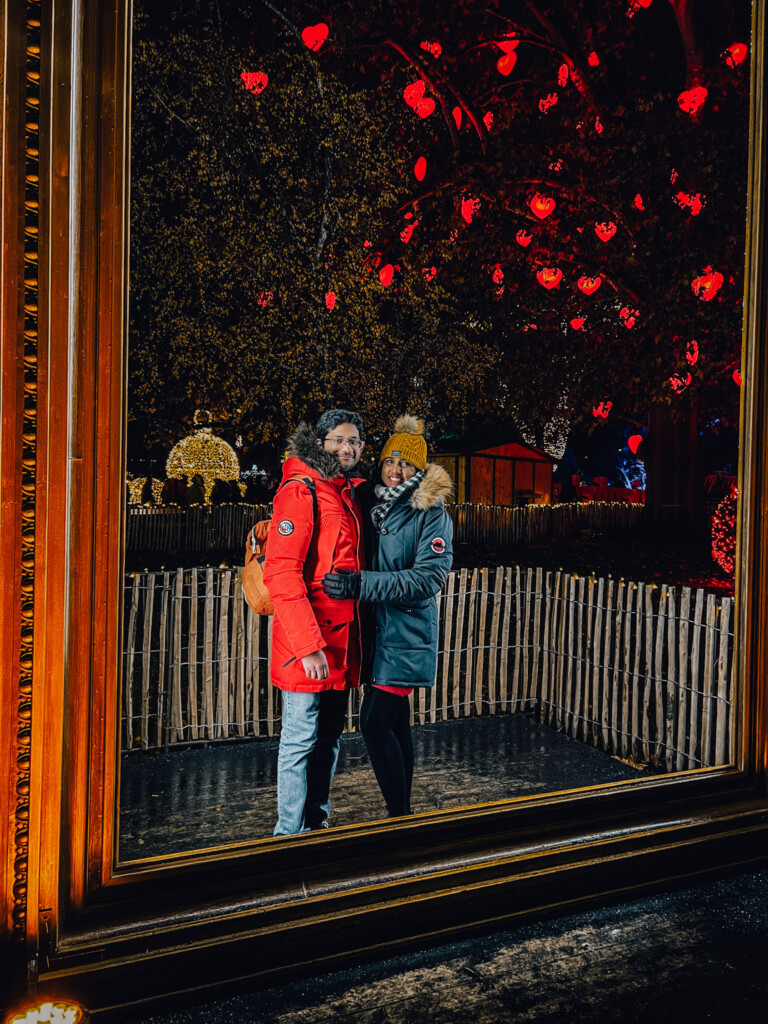 Roopesh and Kiki from RooKiExplorers posing behind a big photo frame with the tree of hearts decoration in the background in Rathausplatz Christkindlmarkt, Vienna.