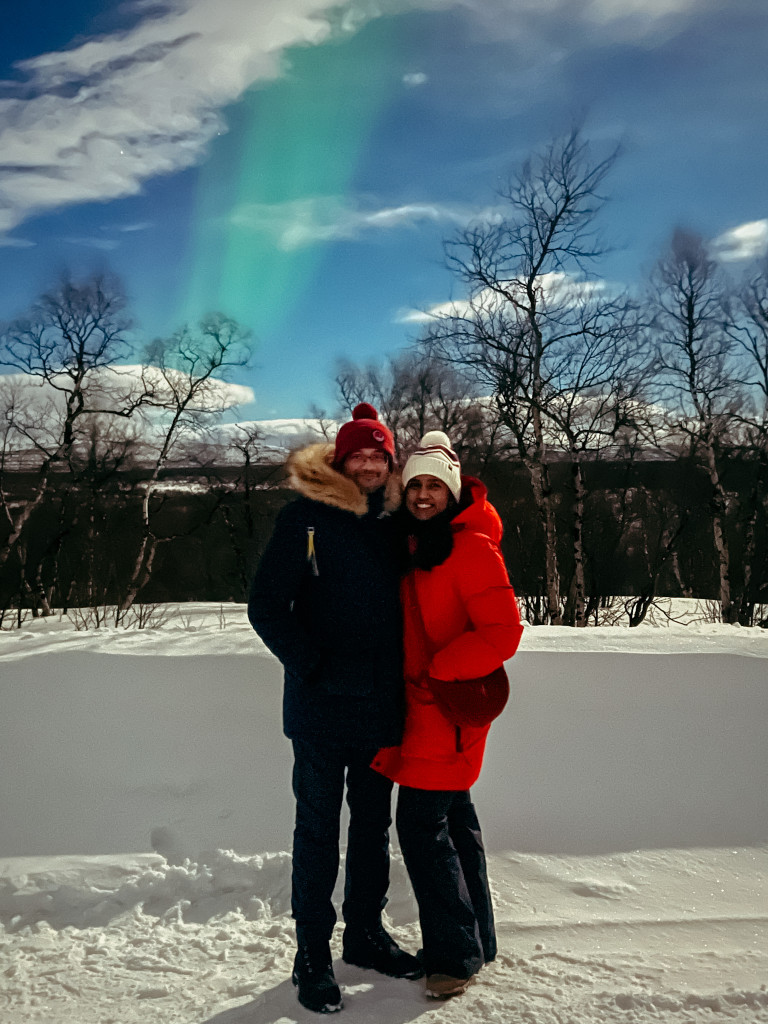 Roopesh and Kiki from RooKiExplorers posing in front of the Northern Lights in Sweden.